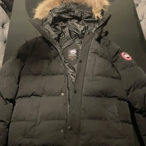 Svart dunjacka med pälsdetalj - Säljer en svart dunjacka från Canada Goose i mycket bra skick. Jackan har en avtagbar huva med pälsdetalj och är perfekt för kalla vinterdagar. Den har en dragkedja och knappar framtill samt loggan på armen. Supervarm och stilren! Osäker på äkthet där av priset, som också kan diskuteras