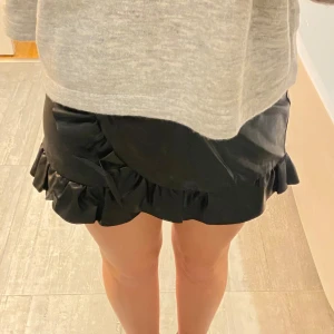 Svart kjol med volang - Superfin svart kortkjol med volangdetaljer. Skinnliknande material, passar perfekt till både vardag och fest!