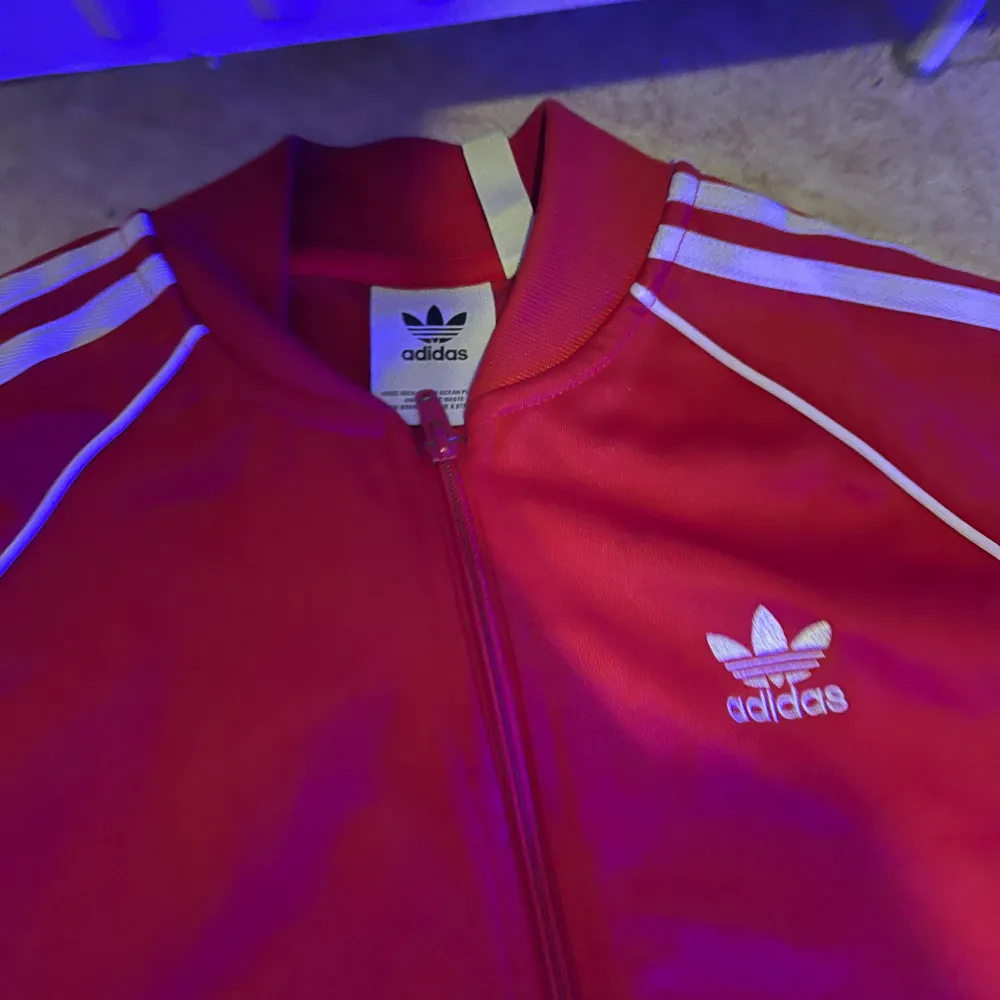 Säljer en snygg röd träningsjacka från Adidas med de klassiska tre vita ränderna längs ärmarna. Jackan har dragkedja och Adidas-loggan på bröstet. Perfekt för träning eller som en sportig vardagslook.. Jackor.