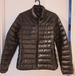 Svart pufferjacka - Säljer en snygg svart pufferjacka i mycket bra skick. Finns dock en liten skada på ärmen som går och dölja med textil märke. Den är perfekt för kyliga dagar och har en dragkedja framtill. Jackan är långärmad och har en glansig finish som ger en modern look. Passar både till vardags och lite finare tillfällen.