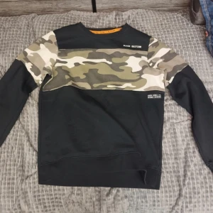 En fin sweatshirt med kamouflage - Säljer en cool svart tröja från Seven Zero med kamouflagemönstrade detaljer på ärmarna.Har användas ett fåtals gånger