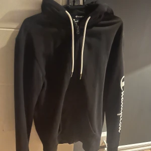 Svart hoodie från Champion - Säljer en svart hoodie från Champion i storlek S. Den har en dragkedja framtill och vita snören vid huvan. Champion-loggan är tryckt på ärmen. Den är i använt skick men ändå bra. 