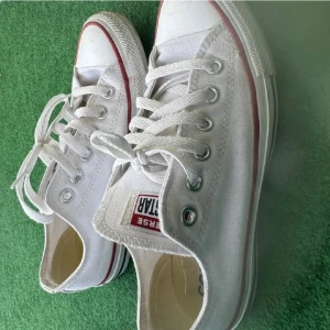 Vita Converse sneakers - Säljer ett par klassiska vita Converse sneakers i bra skick. De har den ikoniska röda och blå randen runt sulan och är perfekta för vardagsbruk. Skorna har snörning och är gjorda i slitstarkt canvasmaterial.
