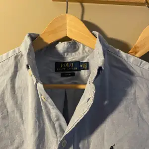 Säljer en klassisk ljusblå skjorta från Polo Ralph Lauren. Skjortan har långa ärmar och knappar framtill. Den är i bomull och har en broderad logga på bröstet. Perfekt för både vardag och fest!