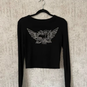 Svart långärmad topp med tryck - Säljer en svart långärmad topp med ett coolt tryck av vingar och rosor på framsidan. Toppen är croppad och har en tight passform, perfekt för en avslappnad men stilren look.