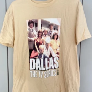 Beige T-shirt med Dallas TV-serie tryck - Säljer en beige T-shirt med ett stort tryck av Dallas TV-serien på framsidan. Perfekt för alla fans av den klassiska serien! T-shirten är kortärmad och gjord i ett mjukt material som känns skönt mot huden.
