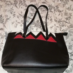 Svart axelväska med röd och vit detalj från Sasha - Säljer en snygg svart axelväska från Sasha med coola röda och vita triangulära detaljer på framsidan. Väskan har långa axelremmar och en dragkedja för säker förvaring. Insidan är fodrad med ett ljust tyg och har en praktisk innerficka med dragkedja. Perfekt för både vardag och fest!