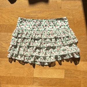 Volangkjol  - Mönstrad volangkjol med små jordgubbar på. Har en dragkedja på sidan. Materialet vid midjan är satin. Står storlek M på lappen men skulle säga att det är XS-S Midja: 36cm Längd: 31cm