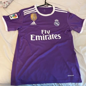 Lila Real Madrid fotbollströja - Säljer en snygg lila Real Madrid fotbollströja med 'Fly Emirates' tryck på framsidan. Tröjan har korta ärmar och är i Climacool-material för bra ventilation. Den har också Real Madrids klubbmärke och LFP-logga på ärmen. På baksidan står det 'Ronaldo 7'. Perfekt för alla fotbollsfans! Säljer för 320kr! 