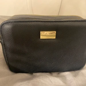 Svart axelväska med dragkedja - Säljer en svart axelväska i skinn med gulddetaljer. Väskan har två dragkedjor upptill och en innerficka med dragkedja. köpt från Åhléns, axelbanden kan man ta av🤗