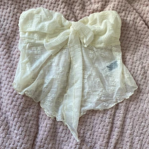 Beige axelbandslös topp från Cider - Säljer en superfin beige axelbandslös topp från Cider. Den är i ett lätt och luftigt material med en söt rosett framtill. Perfekt för sommarens fester eller en dag på stan. Toppen är i nyskick och har en romantisk känsla med sitt spetsliknande tyg.
