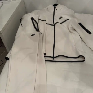 Vit jacka från Nike - Säljer en snygg vit jacka från Nike med svarta detaljer. Den har en dragkedja framtill och är långärmad. Perfekt för träning eller vardagsbruk. Jackan är i nyskick och har en sportig stil.