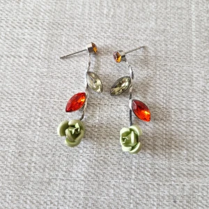 Färgglada örhängen med blommor och stenar - Säljer ett par unika dinglande örhängen. De har en silverfärgad bas med två glittriga stenar i rött och grönt. Längst ner finns en söt grön blomma som ger en extra touch. Perfekta för att liva upp vilken outfit som helst!