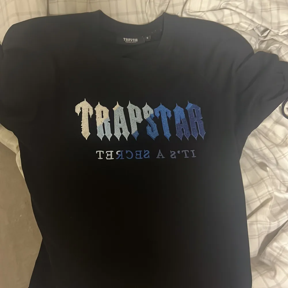 Säljer en svart t-shirt från Trapstar med texten 'TRAPSTAR' i en cool gradient från vit till blå. Texten 'IT'S A SECRET' är tryckt under i mindre bokstäver. T-shirten är kortärmad och i nyskick. Perfekt för en avslappnad stil!. T-shirts.