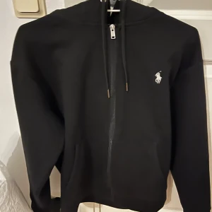 Svart hoodie från Polo Ralph Lauren - Snygg svart hoodie från Polo Ralph Lauren med dragkedja och det klassiska broderade logotypen på bröstet. Perfekt för en stilren och bekväm look. Pris går att diskuteras