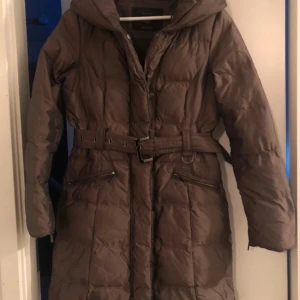 Brun quiltad dunjacka med bälte - Säljer en brun quiltad dunjacka med bälte och huva. Jackan har långa ärmar och dragkedja framtill. Perfekt för kyliga dagar.