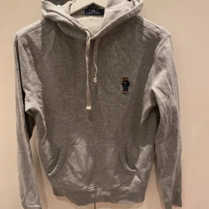 Grå hoodie från Polo Ralph Lauren - Säljer en grå hoodie från Polo Ralph Lauren med en broderad björn på bröstet. Tröjan har en klassisk känguruficka och justerbar huva med snören. Perfekt för en avslappnad stil.