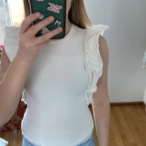 Vit topp med volangdetaljer från Gina Tricot - Säljer en söt vit topp från Gina Tricot i storlek XXS. Toppen har volangdetaljer längs axlarna och är ärmlös. Perfekt för en somrig look!