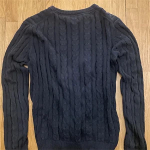 Mörkblå stickad tröja med emblem - Säljer en mörkblå stickad tröja med ett diskret emblem på bröstet. Tröjan har ett klassiskt flätmönster och långa ärmar. Perfekt för kyliga dagar.