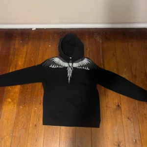Svart hoodie från Marcelo Burlon - Snygg svart hoodie från Marcelo Burlon med ett unikt vingmönster i vitt och grått över axlarna. Perfekt för en stilren och modern look. Perfekt för en lite mer avslappnad och stilig vardag.