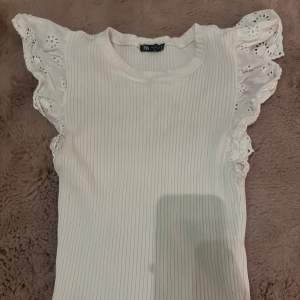 Säljer en söt vit ribbad topp från Zara med spetsdetaljer på ärmarna. Toppen har en rund halsringning och korta ärmar med volang. Perfekt för en somrig look!