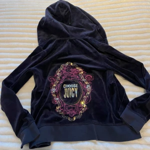Mörkblå velour hoodie från Juicy Couture - Snygg mörkblå velour hoodie från Juicy Couture med dragkedja och huva. På ryggen finns en dekorativ broderad design med texten 'Choose Juicy' i glittriga detaljer. Perfekt för en avslappnad stil.
