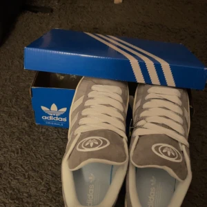 Grå och vita sneakers från Adidas - Snygga grå och vita sneakers från Adidas med klassisk design. Skorna har en överdel i mocka och vita snören. De kommer i originalkartong och har en unik detalj med en liten figur hängande på sidan. Gråa skosnören tillkommer
