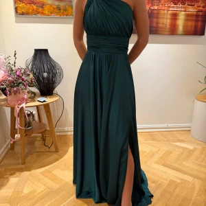 Mörkgrön långklänning med oneshoulder - Elegant mörkgrön långklänning med oneshoulder-design och draperad överdel. Klänningen har en smickrande passform med markerad midja och en stilfull slits framtill. Perfekt för festliga tillfällen. Klänningen är använd en gång på en bal och därför har den lite fläckar på de delar som släppas på marken