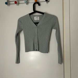 Grå ribbad kofta från Zara - Säljer en stilren grå ribbad kofta från Zara. Den har en v-ringad design med knappar framtill och långa ärmar. Perfekt för lager-på-lager under kyligare dagar. Den är lite mer åt det mint färgade hållet