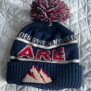 Blå mössa med tofs och Åre-broderi - Snygg blå mössa med stor tofs i rött, vitt och blått. Broderad text 'Åre' och bergsmotiv framtill. Perfekt för kalla dagar!