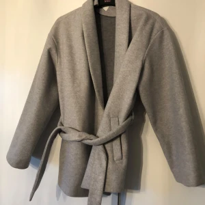 Grå kappa med knytband - Elegant grå kappa från Vila med knytband i midjan. Den har en stilren design med långa ärmar och en mjuk, bekväm passform. Perfekt för kyligare dagar.