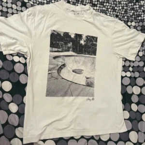 Vit t-shirt med skateboardmotiv - Vit t-shirt med ett stort svartvitt tryck av en skateboardramp. Perfekt för skate-entusiaster som vill ha en stilren look.