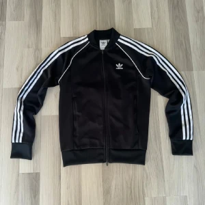 Svart träningsjacka från Adidas - Snygg svart träningsjacka från Adidas med de klassiska tre vita ränderna längs ärmarna. Jackan har dragkedja framtill och är långärmad med ribbade muddar. Perfekt för sportiga aktiviteter eller en avslappnad stil. Storlek S