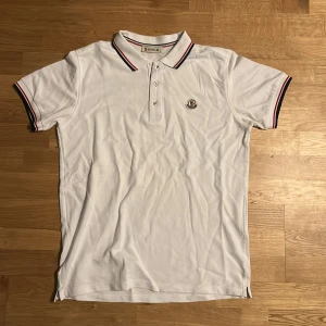 Vit pikétröja från Moncler - Säljer en stilren vit pikétröja från Moncler med klassisk krage och knappar. Tröjan har röd och blå randdetalj på krage och ärmslut samt Moncler-logga på bröstet. Perfekt för en avslappnad men snygg look inför sommaren och våren.