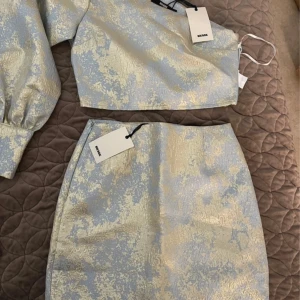 Blå och guld topp och kjol från Bik Bok - Snyggt set med en kort topp och kjol i ett glansigt blått och guldigt mönster från Bik Bok. Toppen har långa ärmar och en elegant struktur. Perfekt för en stilfull look. Storlek S i topp och xs i kjol. Helt ny med prislapp