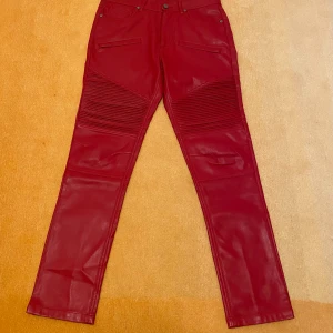 Röda skinnbyxor från Zara - Snygga röda skinnbyxor i imitation från Zara med coola ribbade detaljer på låren. Byxorna har en högmidjad passform och stängs med dragkedja och knapp. Perfekta för att ge din outfit en edgy touch. Se bilder för mått!