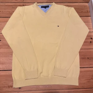 Gul tröja från Tommy Hilfiger - Säljer en stilren gul tröja från Tommy Hilfiger med v-ringning och ribbade muddar. Perfekt för en avslappnad och snygg look. Den har en liten logotyp på bröstet.