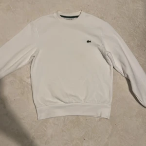 Vit sweatshirt från Lacoste - Säljer en klassisk vit sweatshirt från Lacoste med deras ikoniska logga på bröstet. Tröjan har en normal passform och är perfekt för en stilren look. Den är långärmad och tillverkad i ett mjukt material för extra komfort.