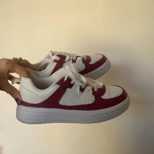 Vita och Vinröda sneakers - Snygga vita sneakers med röda detaljer och snörning. Perfekta för en casual look. Skorna har en klassisk design med perforerade detaljer på sidorna. Storlek 38 men sitter som 37, därför säljs de tyvärr. Väldigt lätta och använda en gång ✨