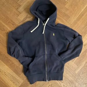 Mörkblå hoodie från Ralph Lauren - Säljer en snygg mörkblå hoodie från Ralph Lauren med dragkedja och vit snörning i luvan. Den har en liten gul logga på bröstet och är perfekt för en avslappnad stil. Passar bra till både jeans och träningsbyxor. Står M men passar S. Passar lite litet på mig som är 186