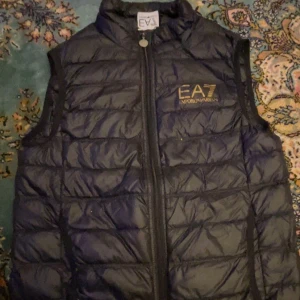 Svart dunväst  EA7 - Snygg svart dunväst från EA7 med dragkedja framtill. Perfekt för kyligare dagar och har en stilren design med märkets logga på bröstet.