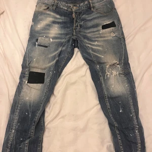 Blå jeans från Dsquared2 - Säljer ett par blå jeans från Dsquared2 med slitna detaljer och färgstänk. Byxorna har en straight passform och är tillverkade i denim. De har en klassisk femficksdesign och en läderpatch med logotyp baktill.
