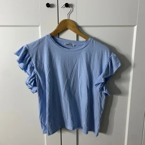 Blå topp med volangärmar från Zara - Säljer en ljusblå topp från Zara med volangdetaljer på ärmarna. Perfekt för en somrig look. Toppen har en rund halsringning och är tillverkad i ett mjukt material.