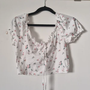 Vit blommig croppad topp - Charmig vit croppad topp med blommigt mönster i rosa och grönt. Toppen har korta ärmar med volangdetaljer och en söt knytning framtill. Perfekt för en somrig look!
