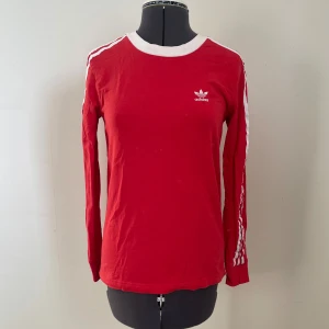 Röd långärmad tröja från Adidas - Snygg röd långärmad tröja från Adidas med vita ränder längs ärmarna och logga på bröstet. 