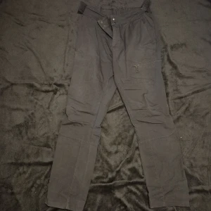 Svarta överdragsbyxor med dragkedja - Säljer ett par svarta överdragsbyxor med praktiska fickor och dragkedja. Byxorna har en avslappnad passform och är perfekta för en casual look. De är tillverkade i ett slitstarkt material och har knappar i midjan.