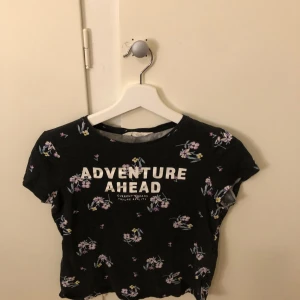 Svart blommig topp från H&M - Säljer en svart topp från H&M med texten 'ADVENTURE AHEAD' i vitt. Toppen har ett blommigt mönster i lila och rosa samt korta ärmar. Perfekt för en avslappnad stil.