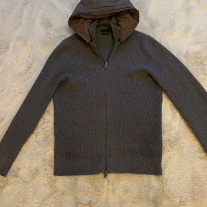 Grå stickad cardigan från Massimo Dutti - Säljer en stilren grå stickad jacka från Massimo Dutti i extrafin ull. Jackan har en praktisk dragkedja och en quiltad huva för extra värme. Perfekt för kyliga dagar.