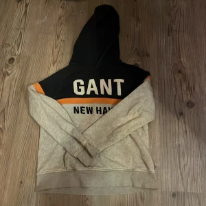 Beige och svart hoodie från GANT - Säljer en snygg hoodie från GANT i beige och svart med orange detaljer. Tröjan har en stor GANT-logga på bröstet och en bekväm huva. Perfekt för en avslappnad stil. Den har några prickar på backsidan men det går någ bort om man tvättar den rijält 