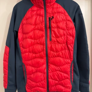 Röd och marinblå jacka från Peak Performance - Snygg och funktionell jacka från Peak Performance i röd och marinblå färg. Jackan har en quiltad design med dragkedja och en praktisk huva. Perfekt för kyligare dagar.